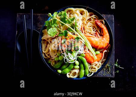Ciotola colorata di ramen con gamberi, edamame, gnocchi, spaghetti ed erbe fresche in brodo, con bacchette su sfondo scuro Foto Stock