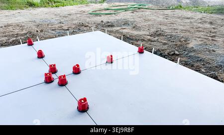 Fondazioni per solette in calcestruzzo con distanziali di livellamento rossi posizionati su una superficie di ghiaia, circondate da erba e un tubo flessibile da giardino sullo sfondo Foto Stock