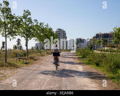 Donna in bicicletta su un sentiero sterrato con edifici urbani e alberi sullo sfondo Foto Stock