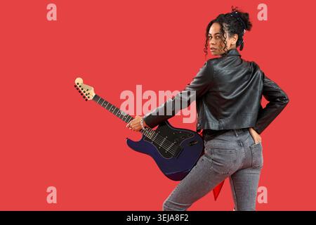 Elegante giovane donna afroamericana in giacca di pelle con chitarra elettrica su sfondo rosso Foto Stock