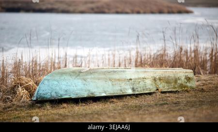La barca turchese intemprata si appoggia capovolta su una riva erbosa. Alte canne secche fiancheggiano il tranquillo lago ghiacciato sotto il cielo coperto. Tranquillo e rustico paesaggio invernale Foto Stock