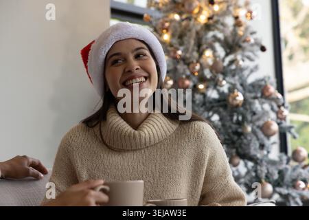 Amici seduti sul divano accanto all'albero di Natale floccato, donna asiatica che indossa un maglione beige che tiene la tazza Foto Stock
