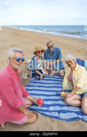 Amici anziani che si riuniscono su una coperta a strisce blu sulla spiaggia di sabbia, gustando drink e chiacchierando, incarnando l'invecchiamento attivo e la connessione sociale Foto Stock