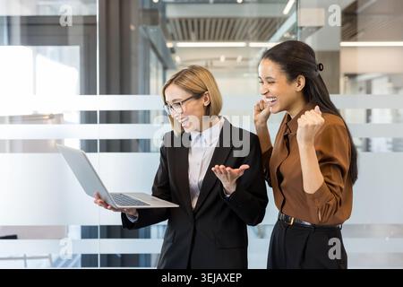 Due diverse donne d'affari celebrano con entusiasmo un risultato di successo, stringendo pugni e ridendo gioiosamente mentre guardano le informazioni su un notebook in un ufficio moderno Foto Stock