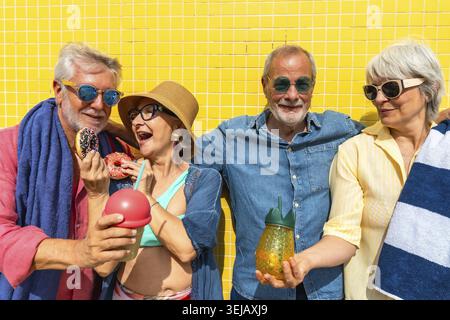 Un gruppo di diversi amici anziani che ridono e sorridono mentre tengono in mano ciambelle colorate e bevande rinfrescanti, celebrando l'amicizia contro un urlo luminoso Foto Stock