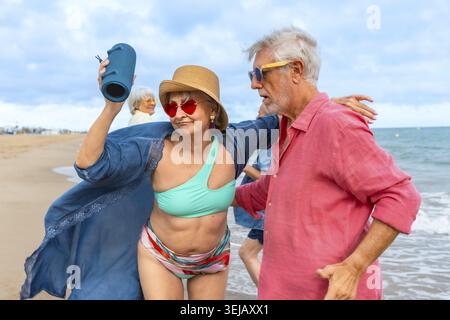 Amici anziani che ballano e ascoltano musica da un altoparlante portatile su una spiaggia soleggiata, si godono la pensione attiva, le risate, le onde e il summ spensierato Foto Stock