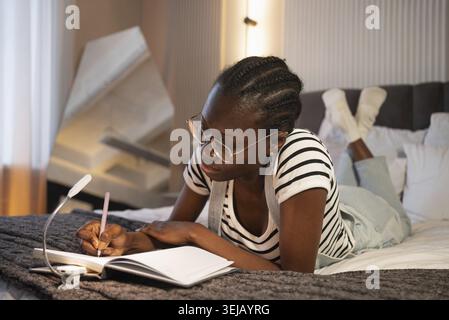 Giovane donna afro-americana che indossa gli occhiali, sdraiata comodamente sul letto, scrivendo con cura in un notebook mentre utilizza una lampada a clip per enha Foto Stock