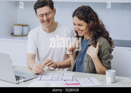 Una coppia sorridente sta lavorando a un nuovo progetto di appartamento, con un computer portatile e documenti con disegni della casa sparsi sul tavolo. Casa di famiglia Foto Stock