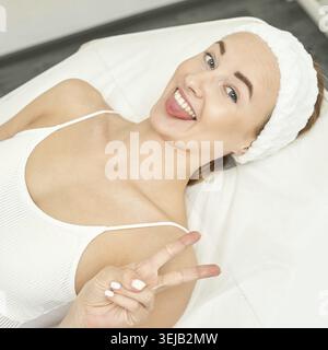Giovane donna caucasica con fascia sorridente e segno di pace mentre sdraiata sul tavolo spa Foto Stock