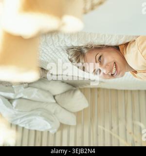 Giovane donna caucasica che si rilassa sul letto sorridendo in alto piacevolmente in una camera da letto luminosa e accogliente Foto Stock