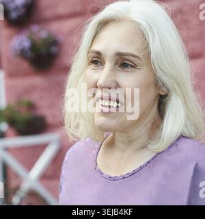 Anziana donna caucasica sorridente in lavanda con sfondo rosa e dettagli floreali Foto Stock