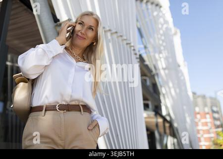 Una donna con lunghi capelli biondi sorride mentre parla al telefono. È vestita con una camicetta bianca e pantaloni beige, in piedi vicino a un elegante e truffatore Foto Stock