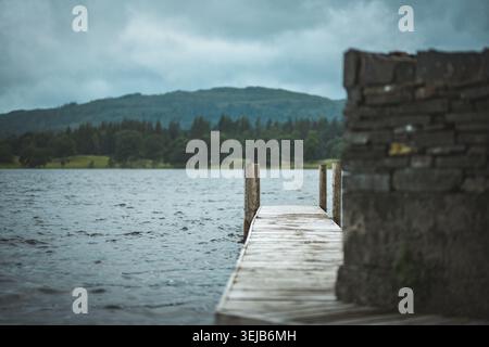 Un molo di legno intemprato si estende in un lago calmo sotto un cielo umido e coperto, con una struttura in pietra sulla destra e colline boscose in lontananza Foto Stock
