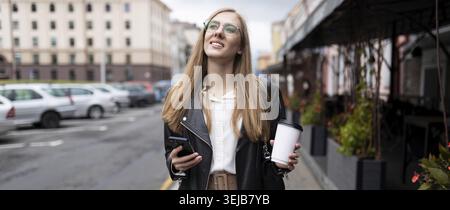 Giovane donna forte che cammina per la città con una tazza di caffè in mano guarda in lontananza Foto Stock