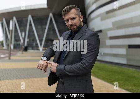 Un uomo adulto con una tuta da lavoro punta il dito verso l'orologio sullo sfondo di un edificio per uffici Foto Stock