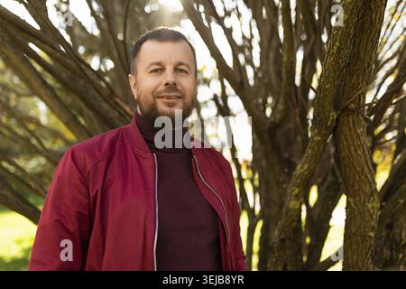Giovane sorridente con barba in giacca rossa nel parco autunnale Foto Stock