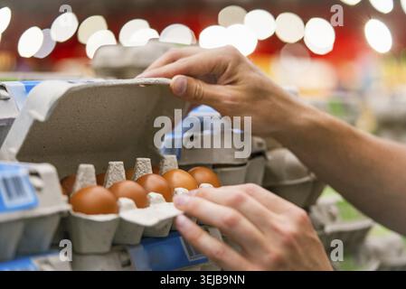 L'uomo controlla con mano un cartone di uova marroni nel corridoio del supermercato, garantendo qualità e freschezza. Selezione di prodotti di qualità nel negozio Foto Stock