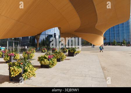 HELSINKI, FINLANDIA - 11 AGOSTO 2025: Baldacchino in legno dorato curvo di un moderno edificio pubblico con piantagioni di fiori in una giornata estiva. Foto Stock