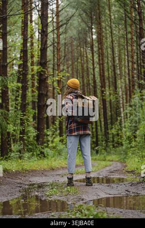 Una giovane escursionista che indossa uno zaino e un berretto giallo si sta godendo una tranquilla passeggiata su un sentiero fangoso della foresta dopo la pioggia, circondato da un lussureggiante tr verde Foto Stock