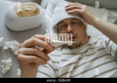 Uomo malato affetto da raffreddore o influenza, sdraiato a letto con un asciugamano bagnato sulla fronte, controllando la temperatura corporea con un termometro, una scatola di tessuti e usato Foto Stock