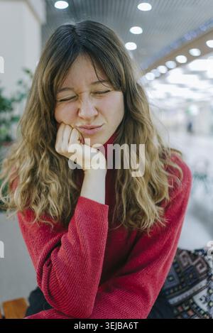 La giovane donna con lunghi capelli ondulati e con un maglione dolcevita rosso è seduta con gli occhi chiusi, mentre si prepara un volto tenendo la testa in mano Foto Stock