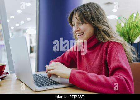 Felice lavoratrice freelance che lavora su un computer portatile in un accogliente bar, gustando una tazza di caffè mentre si gode l'atmosfera rilassata e la luce naturale che scorre Foto Stock