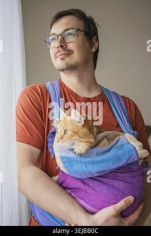 Uomo che indossa una t-shirt arancione e occhiali che tiene in mano il suo amato gatto allo zenzero arancione comodamente accoccolato in un elegante trasportino in tessuto all'interno di una casa, sh Foto Stock