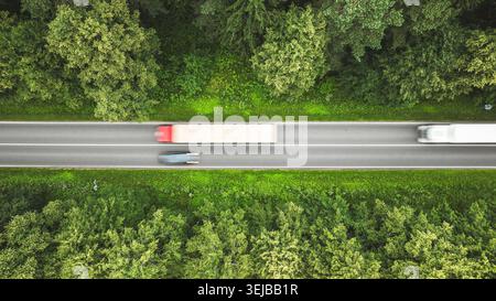 Lo scatto con droni dall'alto verso il basso cattura la sfocatura del movimento di due camion che viaggiano su una strada circondata da fitti boschi, mettendo in risalto i trasporti e la natura Foto Stock