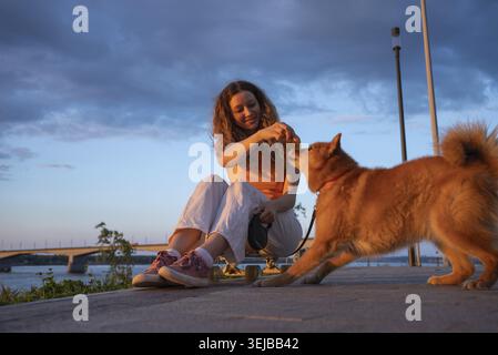 La giovane donna ama trascorrere un po' di tempo con il suo cane da orso careliano, premiandola con una delizia mentre si rilassano sul fiume al tramonto, creando un'emozione sc Foto Stock