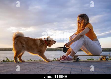 La giovane donna con i capelli lunghi è seduta su uno skateboard vicino al fiume al tramonto, tenendo il guinzaglio del suo cane careliano, godendosi la bugia dell'ora d'oro Foto Stock