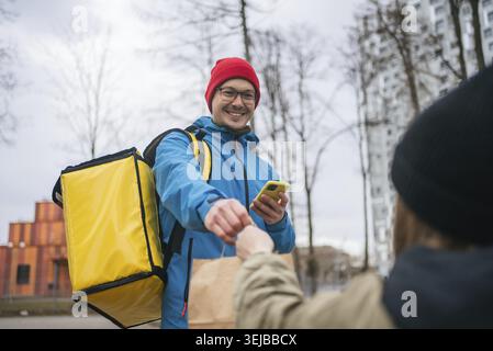 Un addetto alle consegne sorridente che utilizza un'app mobile, consegna un ordine alimentare in uno zaino termico e consegna un sacchetto di carta a un cliente all'aperto in una città Foto Stock