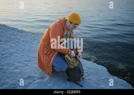 Giovane uomo che indossa una giacca arancione e un berretto giallo aprendo lo zaino mentre si inginocchia sulla riva di un lago ghiacciato al tramonto, godendosi la pace e la tranquillità Foto Stock