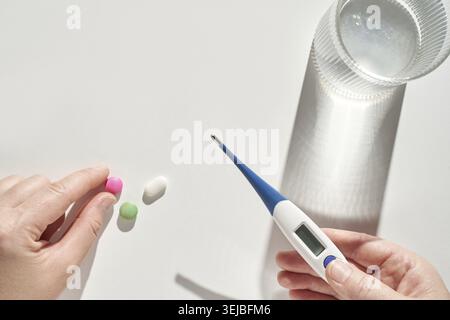 Mani che trattengono pillole e termometro digitale su superficie bianca con un bicchiere d'acqua alla luce del sole Foto Stock