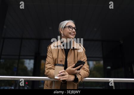 Una donna anziana matura al telefono e una tazza di caffè in mano guarda in lontananza fuori Foto Stock
