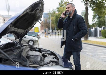 Un uomo adulto dai capelli grigi con barba parla su un telefono cellulare vicino al cofano aperto di un'auto rotta Foto Stock