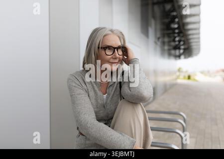 Una donna matura con capelli e occhiali grigi si siede comodamente su un edificio elegante. Guarda l'ambiente circostante, riflettendo in un momento sereno. Il Foto Stock