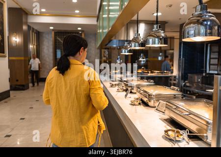 Una donna con lunghi capelli neri che indossa una giacca gialla si trova a un buffet in un hotel. L'ambiente e' moderno con varie opzioni gastronomiche. Foto Stock