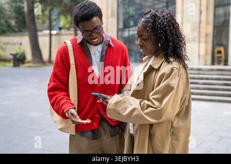 Allegra donna afroamericana sorridente che mostra foto su smartphone a un amico maschio nero positivo. Foto Stock