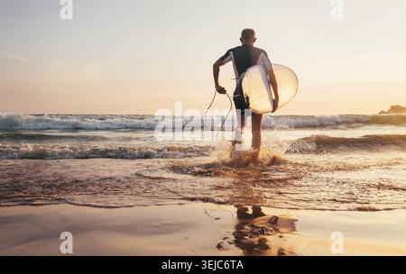 Surfista che tiene una tavola da surf che scorre nell'acqua dell'oceano sulla spiaggia di sabbia durante il tramonto dorato in Sri Lanka, rappresentando lo stile di vita attivo sport acquatici tropicali Foto Stock
