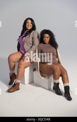 Diverse amiche che posano su cubi bianchi in uno studio professionale, indossando tute e stivali Foto Stock