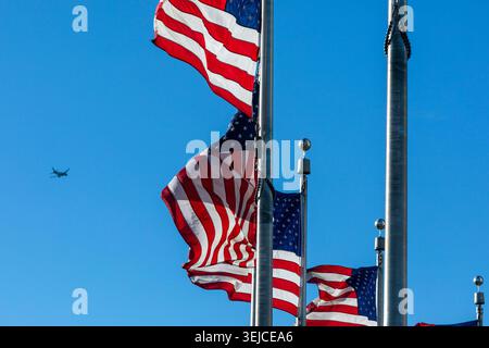 Bandiere americane che sventolano su pali intorno al Washington Monument contro un cielo blu chiaro con un aereo in lontananza in una giornata di sole, Washington DC, USA Foto Stock