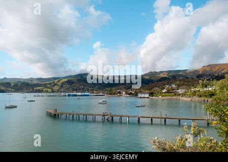 Akaroa, nuova Zelanda: Lungo molo in legno si estende fino all'acqua della baia del porto turchese sotto verdi colline vulcaniche e nuvole. Foto Stock