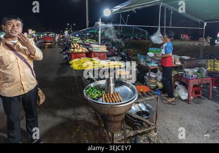 Un venditore e un uomo sono in piedi a uno stand di street food con spiedini su un grill a carbone e prodotti freschi in un vivace mercato notturno a Siem Reap, Cambogia Foto Stock