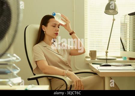 Giovane donna d'affari caucasica che raffredda la fronte con una bottiglia d'acqua seduto alla scrivania dell'ufficio, sperimentando calore stress e stanchezza. Utile per il benessere sul posto di lavoro o per la produttività estiva Foto Stock