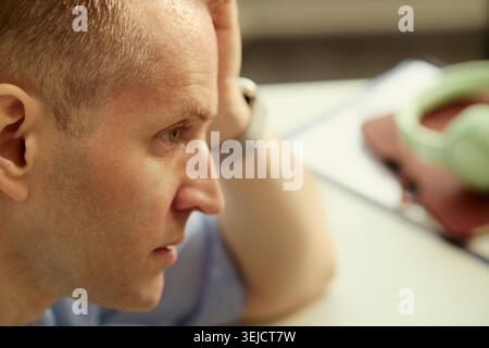 Primo piano di un giovane adulto caucasico che tiene la testa e guarda in basso, mostrando stress e sovraccarico mentale. Utile per burnout, ansia, pressione sul posto di lavoro, campagne di salute mentale Foto Stock