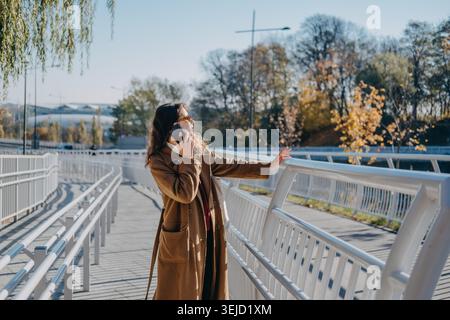 Una donna sta in piedi su un ponte pedonale mentre parla al telefono cellulare appoggiandosi contro le ringhiere. Negoziazione client lavoro a distanza, rete cellulare urbana Foto Stock