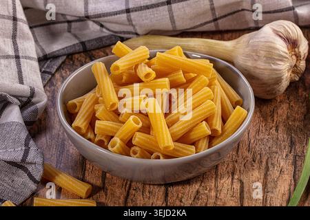 Immagine STILL Life con pasta corta dorata cruda in ciotola grigia, accompagnata da bulbo d'aglio intero, gambo di cipolla verde. Superficie rustica in legno, plaid Foto Stock