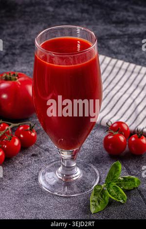 Bicchiere di vivace succo di pomodoro rosso circondato da pomodori freschi maturi, pomodori ciliegini sulla vite, foglie di basilico verde. Bevanda sana su tessuto scuro Foto Stock