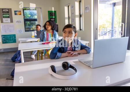 Diversi compagni di classe seduti in classe, bambini con laptop e cuffie, uno con giacca in denim Foto Stock