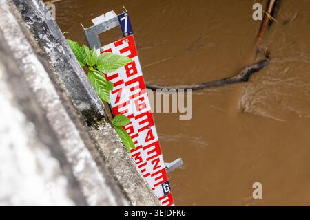 Vista ravvicinata dell'indicatore di livello dell'acqua rosso e bianco che misura l'acqua profonda e fangosa del fiume accanto alla parete di cemento, causando allarmanti Foto Stock